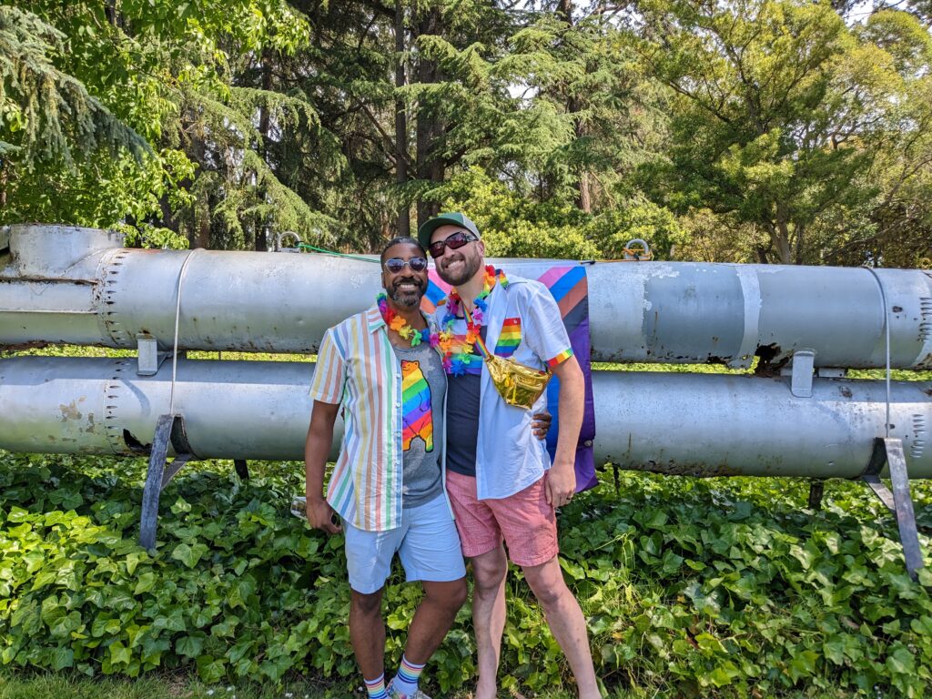 Matt and Bryan in rainbow gear in Alden Park, standing in front of a stacked pair of metal torpedo tubes.